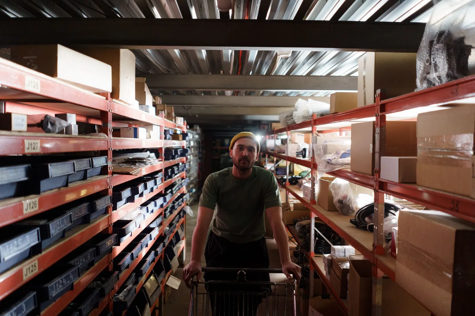 A warehouse worker in a green T-shirt and yellow beanie pushes a wire trolley down a narrow aisle between racking full of bins and boxes.